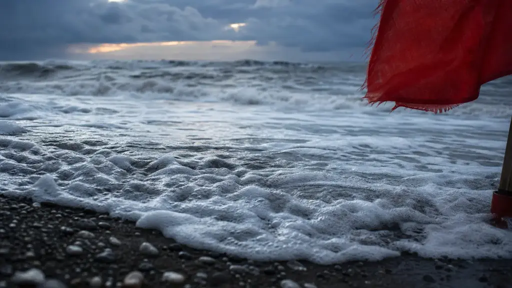 playa de sagunto: espuma y oleaje sobre la arena mojada bajo un cielo de tormenta con una bandera roja