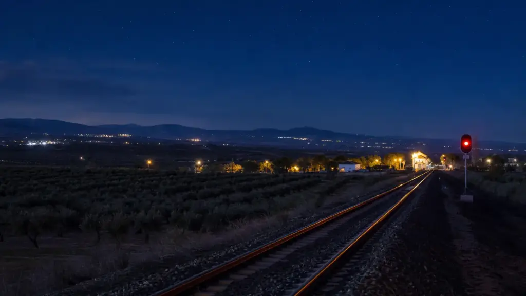 accidente de adamuz: vía de tren de noche entre olivares con una estación iluminada a lo lejos