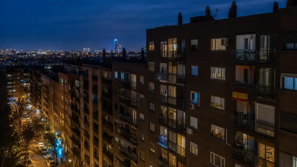 vivienda en españa: edificios de viviendas de noche con ventanas iluminadas y una bandera de España en un balcón