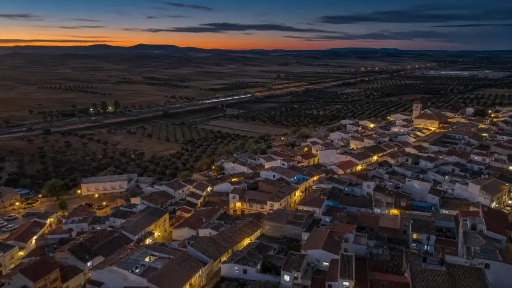 audiencias de horizonte: vista aérea de un pueblo al anochecer con ventanas iluminadas por televisores y campos de olivos